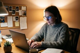 Femme blogueuse portant des lunettes anti-lumière bleue en train d'écrire sur un ordinateur portable le soir dans un bureau à domicile confortable